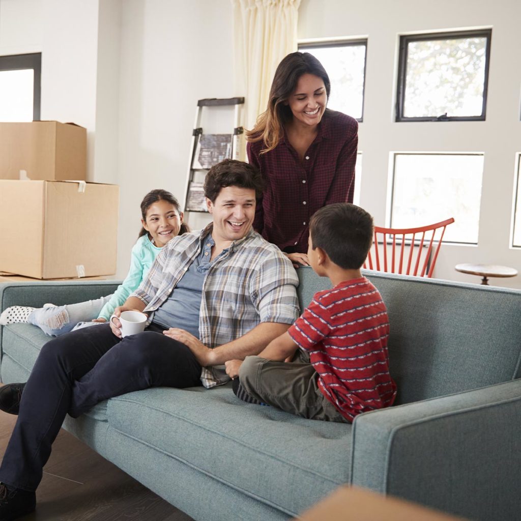 The image captures a joyful family moment inside a home during what appears to be a moving or unpacking process. A man, woman, and two children are gathered around a teal sofa, smiling and talking to each other. The man is seated on the couch, holding a white coffee mug, while the young boy, wearing a red striped shirt, is sitting next to him, engaged in conversation. The girl, sitting at the other end of the couch, smiles warmly. The woman, standing behind the sofa, leans forward with a friendly expression, joining the conversation. In the background, several moving boxes are visible, indicating that the family may have recently moved into the home or is in the process of unpacking. A ladder and some other unpacked items can also be seen, further suggesting a settling-in period. The large windows allow natural light to brighten the room, contributing to a warm and welcoming atmosphere.