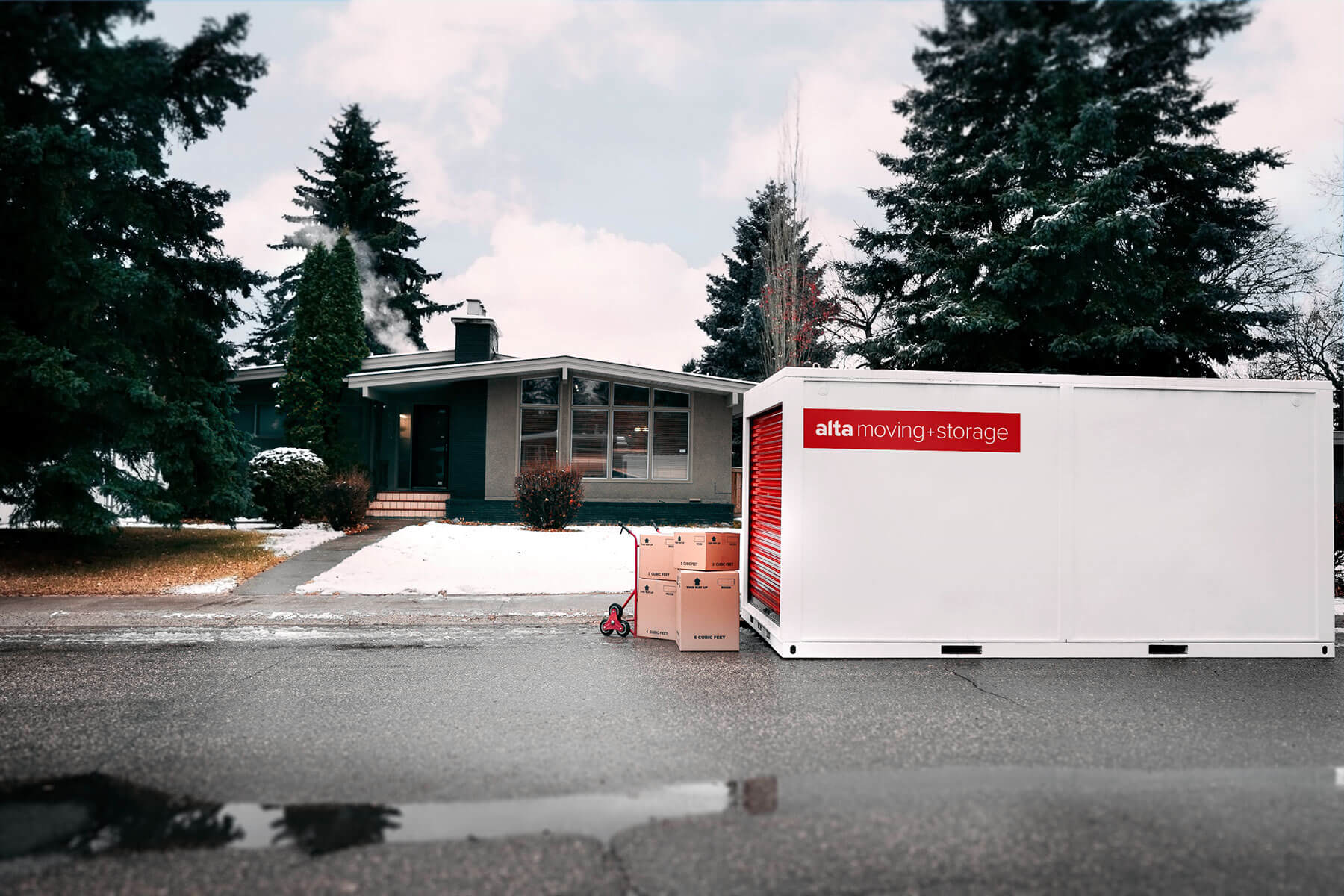 The image shows a suburban house with a portable storage by ALTA STORAGE unit placed in front of it on the street. The house is a one-story, mid-century modern design with large windows and a slightly sloped roof. A paved walkway leads to the entrance, flanked by bare shrubs and patches of snow on the lawn. There are large evergreen trees surrounding the property, contributing to a quiet, wintery atmosphere. The ground is wet, with a small puddle visible on the road in the foreground. On the right side of the image, a white storage container with the branding "ALTA STORAGE" in bold red and black letters is positioned on the street. The container has a red roll-up door, and in front of it, two brown cardboard moving boxes are stacked on a hand truck. The weather appears cold, with snow scattered on the ground and a cloudy sky overhead.